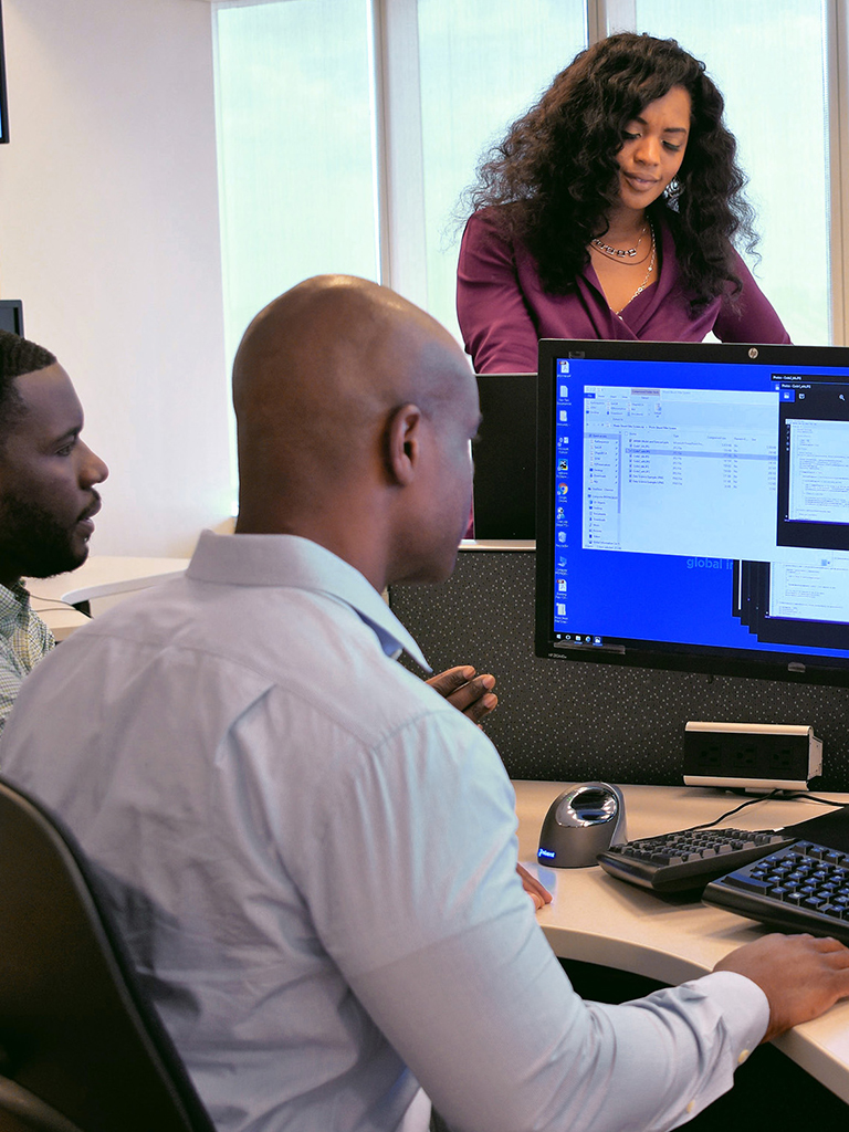 Coworkers sitting at a desk in front of a laptop computer looking at monitors.